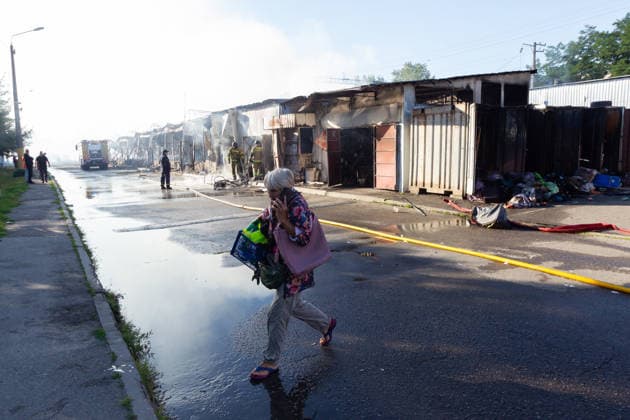 A woman talking on the phone crosses a road as firefighters work at the Antykar market in Obolonskyi district, which caught fire as a result of an overnight Russian missile strike, Kyiv, Ukraine, July 30, 2026. Debris fell on the market near the Pochaina metro station, igniting trading pavilions. The blaze covered an area of about 100 square metres. Traders’ goods were destroyed, leaving only burnt-out metal structures at the site. (Photo by Kyrylo Chubotin Ukrinform) Market in Kyiv catches fire due to overnight Russian attack  Copyright: xKyryloxChubotinx