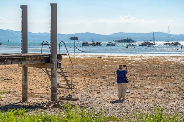 Badesteg ins Trockene, Niedrigwasser am Bodensee, durch Trockenheit freigelegte Uferzone am Bodensee, die Boote liegen weit draußen, die Schifffahrt ist eingeschränkt, Wasserburg, August 2026 Deutschland, Wasserburg, 8. August 2026, Badesteg ins Trockene, Niedrigwasser am Bodensee, durch Trockenheit freigelegte Uferzone am Bodensee, ein Badesteg führt ins Trockene, weit entfernt von der Wasserlinie, eine Besucherin fotografiert die weit zurückgewichene Uferlinie, die aktuelle Trockenheit hat den Pegelstand stark sinken lassen, die Boote liegen weit draußen, die Schifffahrt ist eingeschränkt, Niedrigwasser, Wasserstand sehr niedrig, zu wenig Regen, schönes Wetter, Hochsommer, Hitzewelle mit Temperaturen bis 33 Grad, Sommer, Samstagnac