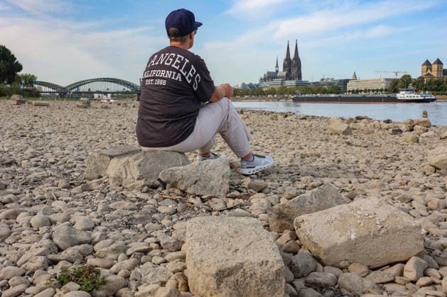 Sommer im Rheinland. Blick auf den Rhein bei Koeln (Köln) - Deutz. Der Rhein in Koeln (Köln) fuehrt (führt) Niedrigwasser aufgrund anhaltender Trockenheit. Im Hintergrund die Hohenzollernbruecke (Hohenzollernbrücke) (links) und der Koelner (Kölner) Dom (rechts). Im Vordergrund sitzt eine Frau auf einem Stein und schaut auf den Rhein. Sommer im Rheinland am 09.08.2026 in Koeln (Köln) Deutschland. *** Summer in the Rhineland: View of the Rhine near Cologne (Deutz). The Rhine in Cologne is at low water levels due to persistent drought. In the background, the Hohenzollern Bridge is on the left and Cologne Cathedral is on the right. In the foreground, a woman is sitting on a rock, looking out at the Rhine. Summer in the Rhineland on Augus