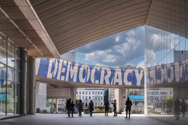 Blick auf den großen blauen Schriftzug DEMOCRACY an der Konrad-Adenauer-Verbindungsbrücke des Europäischen Parlaments. Über dem Brüsseler Europaviertel ziehen im Hintergrund schwere, dunkle Gewitterwolken auf, während sich das Wort verzerrt in der modernen Glasfassade spiegelt. Im Schatten der monumentalen Architektur bewegen sich Passanten als Silhouetten. Die dramatische Lichtstimmung dient als starkes Symbolbild für politische Krisen, die Gefährdung der Demokratie oder gesellschaftliche Spannungen in der Europäischen Union. *** View of the large blue DEMOCRACY signage on the Konrad Adenauer connecting bridge of the European Parliament. In the background, heavy, dark storm clouds gather over the Brussels European Quarter, while the word r GMS20575