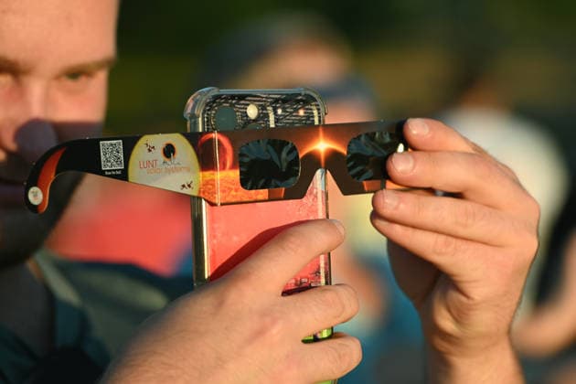 Ein Mann haelt eine Schutzbrille vor sein Smartphone, Menschen beobachten die partielle Sonnenfinsternis und stehen dabei auf der Ripshorstbruecke ueber den Rhein-Herne-Kanal, 12.08.2026 in Oberhausen. *** A man holds a pair of safety goggles in front of his smartphone as people watch the partial solar eclipse while standing on the Ripshorst Bridge over the Rhine-Herne Canal on August 12, 2026, in Oberhausen