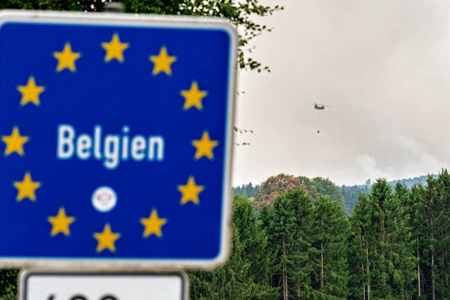 Waldbrand im Hohen Venn Blick von der deutsch-belgischen Grenze bei Kalterherberg - 15.08.26 Blick von Kalterherberg (Gemeinde Monschau) auf den Großbrand im Hohen Venn nahe der deutsch-belgischen Grenze. Im Vordergrund ein blaues Grenzschild im Hintergrund Rauchsäulen des Feuers und ein Löschhubschrauber mit Wasserbehälter im Anflug. Insgesamt zwei Löschhubschrauber sind im Einsatz und werfen jeweils rund 3.000 Liter Wasser ab. Der Hochmoorbrand hat bereits rund 2.000 Hektar Wald- und Moorgebiet erfasst, die tiefen Torfschichten begünstigen einen langanhaltenden Schwelbrand. Auf belgischer Seite laufen bereits Evakuierungen, für Teile Monschaus wird eine mögliche Räumung vorbereitet. Monschau Kalterherberg Nordrhein-Westfalen Copyright: xMarcxJohnx