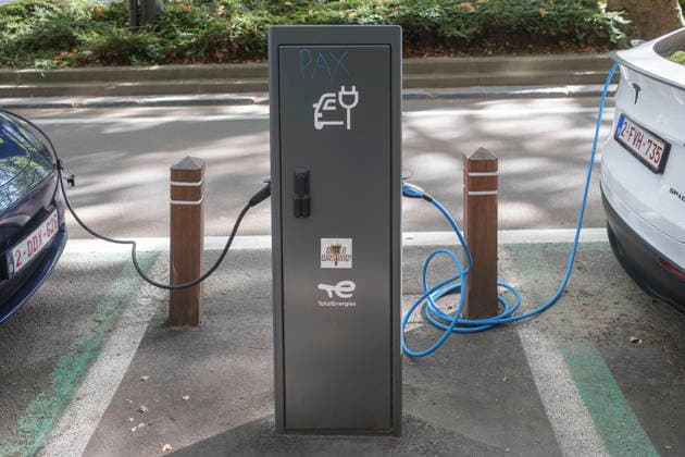 Two electric vehicles are connected via charging cables to an anthracite-colored TotalEnergies charging station at the roadside. On the left, the rear of a blue vehicle is visible, while a white Tesla model with a Belgian license plate charges on the right. In the center, the charging post features the GMS20611