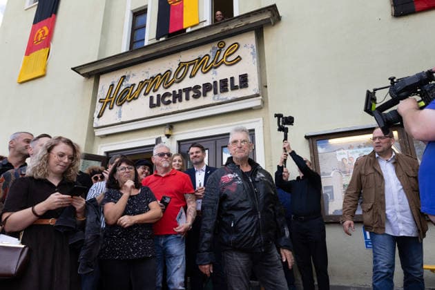 September 5, 2026, movie premiere; Comrade Martin Semmelrogge, actor playing the role of Mr. Glück the pharmacist, wearing a mask, can be seen in the foreground wearing a leather jacket in front of the entrance to the Markneukirchen Harmonie Lichtspiele cinema in Saxony, Germany
