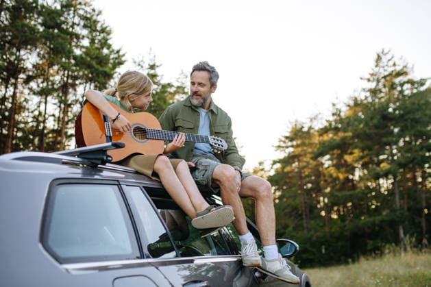 Dad teaching boy how to play guitar during summer camping adventure in nature. Father teaching boy how to play guitar during summer camping adventure sitting on car. model released
