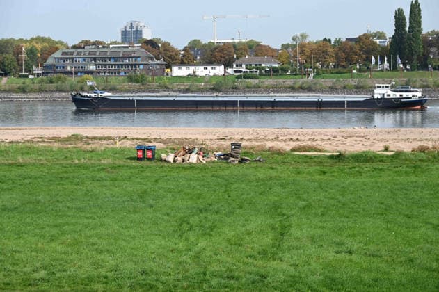 Low water levels in the Rhine have exposed a large sandbar; rocks and sand are visible on the riverbank near the Theodor Heuss Bridge; a cargo ship is sailing in the channel; in the foreground, flotsam, car tires, and debris have been exposed by the low water, Unidentified individuals collected the trash and left it on the bank for pickup; low water levels