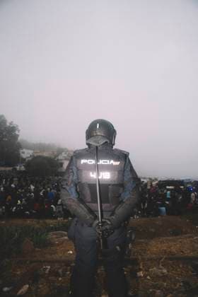 October 4, 2026, Ceuta, Ceuta, Spain: A riot police officer stands guard in the fog in front of migrants sitting on the ground during the eviction of the makeshift camp on El Trampolin beach in Ceuta. Spanish National Police began evicting the makeshift camp on El Trampolin beach in Ceuta on October 4, where more than 3,000 migrants, most of them young Moroccan men, were living. Dozens of police vans, armoured vehicles of the Police Intervention Units (UIP), drones and boats took part in the operation. People were taken by bus to the temporary centre at the Tarajal border to be identified, and the authorities plan to clear the beach in phases before October 15. The camp grew after a mass arrival at the end of July 2026 set off a migration a - ZUMAs392 20261004_zip_s392_002 Copyright: xFabianxSuspensivox