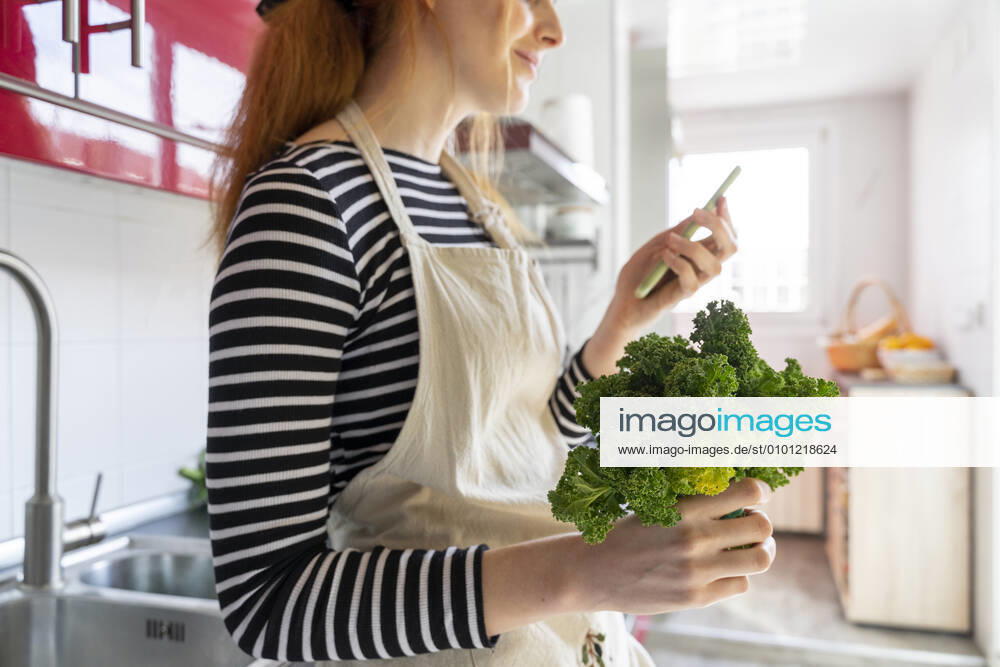 Young woman standing in kitchen, holding cale, using smartphone model released Symbolfoto property released AFVF06038