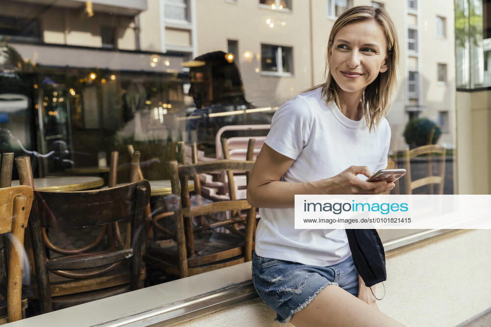 Portrait of woman with smartphone and protective mask in her hand waiting in front of a coffee shop model released Symbolfoto MFF05639
