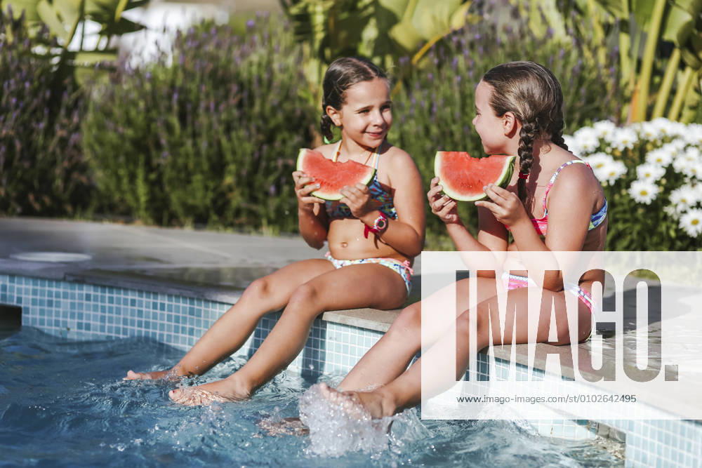 Happy twin girls sitting with watermelon slice at poolside on sunny day model released Symbolfoto property released LJF01574