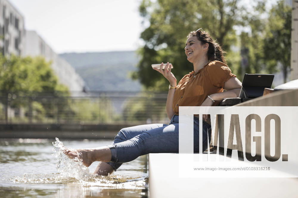 IMAGO Creative: Sommer, Sommertage im Stadtpark Cheerful woman with legs in pond talking over smart phone while sitting at park model released Symbolfoto UUF20794