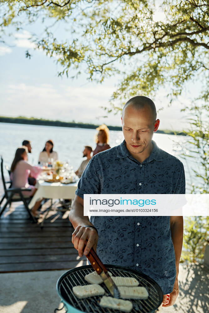 Man grilling Halloumi cheese at a lake with friends in background model released Symbolfoto ZEDF03564