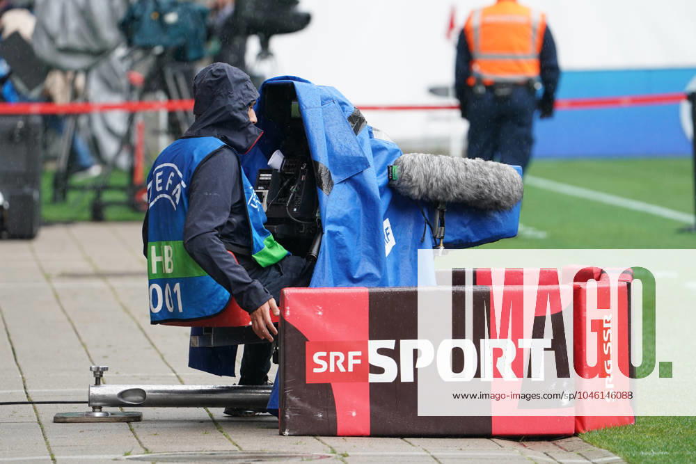 Cameraman at a UEFA event, SRF Sport, SRG SSR, rainy weather, camera with weather protection, sports broadcast, live coverage, soccer match, media coverage, television production, camera technology, sports journalism, security forces in the background, sports event, TV station, broadcasting, Switzerland, stadium, outside broadcast, UEFA HB host broadcaster, sports reporter symbol image