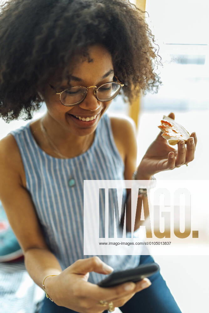 Close-up of smiling businesswoman with curly hair holding bread while using smart phone at home model released Symbolfoto property released BOYF01528