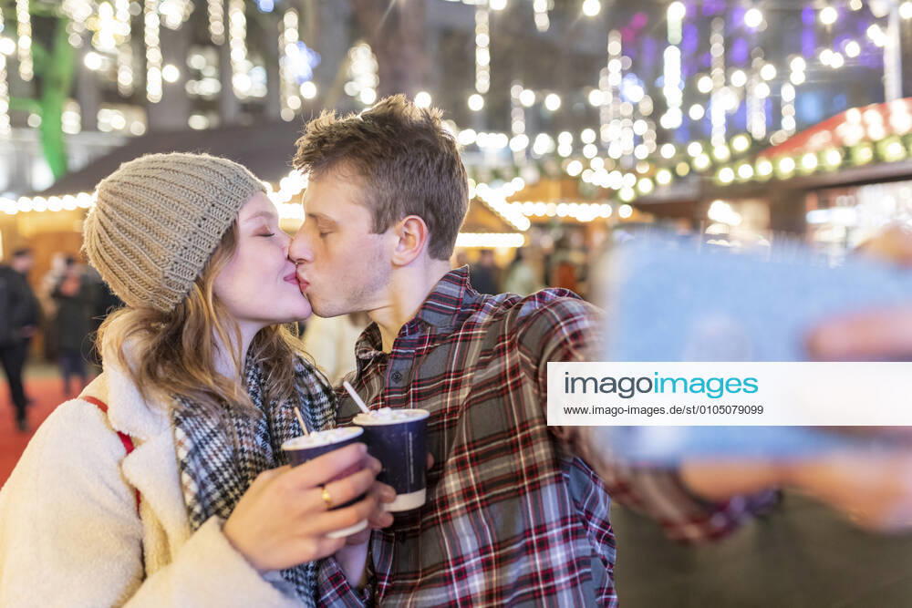 Couple holding hot chocolates kissing while taking selfie in Christmas market at night model released Symbolfoto WPEF03132