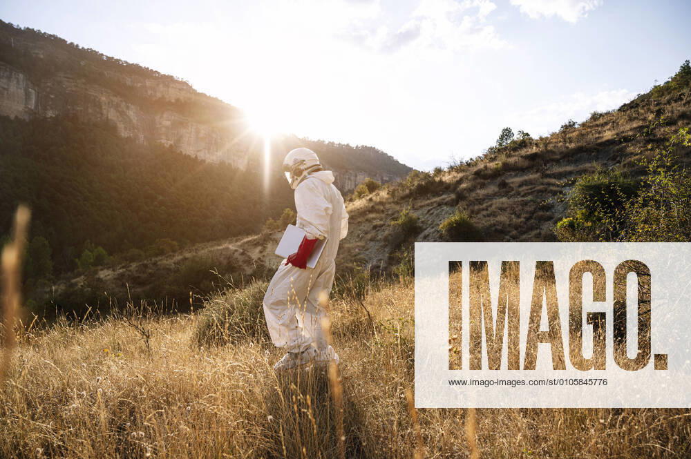 IMAGO Creative: Klimawandel - Zukunft und Innovation Male astronaut with laptop walking on grassy land against sky during sunny day model released Symbolfoto JCMF01438
