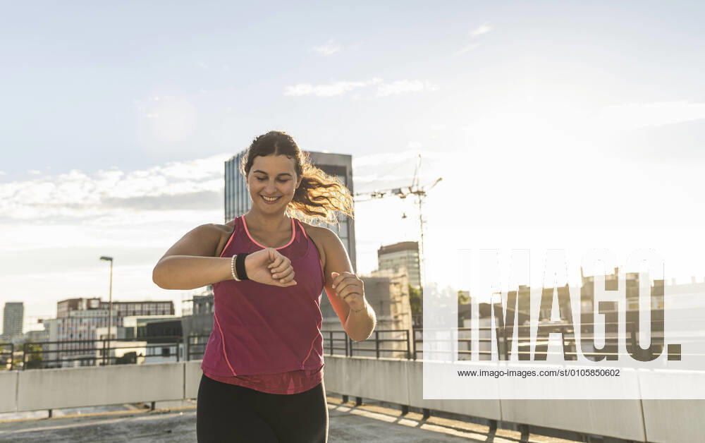 Smiling young woman checking time while running on terrace against sky model released Symbolfoto UUF21378