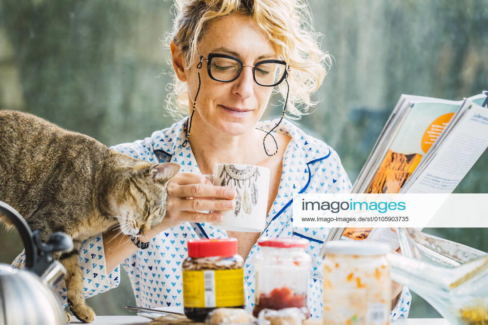 Blonde young mature woman with glasses in pijamas at home in breakfast time, reading a magazine and having a moment with her cat. SPAIN