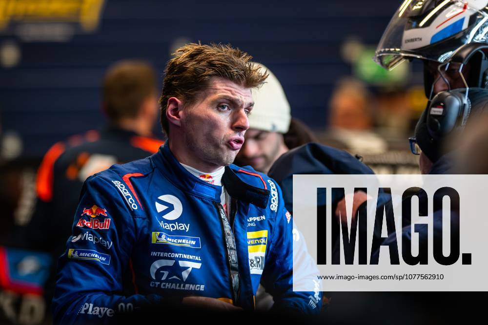 Max Verstappen stands in the paddock wearing his blue racing driver overalls with Red Bull and various sponsor logos The racing driver is focused and talks to team members in the background Max Verstappen in the paddock of the 24h Nürburgring 2026