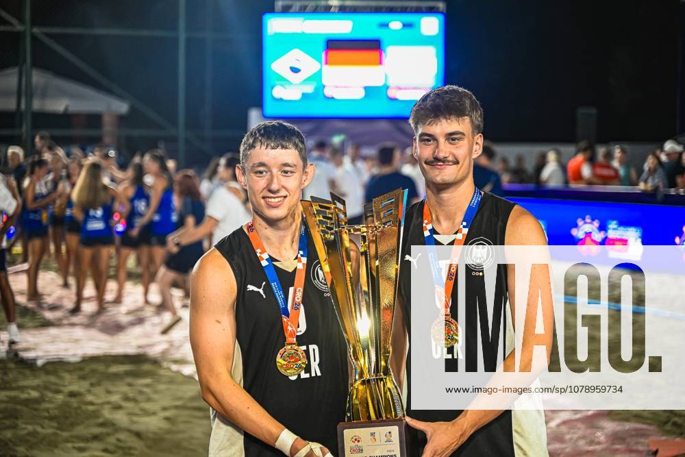 Two players from the German national beach handball team are holding the golden championship trophy together and wearing gold medals around their necks. In the background, the scoreboard with the German flag and celebrating spectators at Lake Jarun in Zagreb can be seen. IHF Beach Handball World Championship 2026, June 23, 2026, Zagreb, Lake Jarun