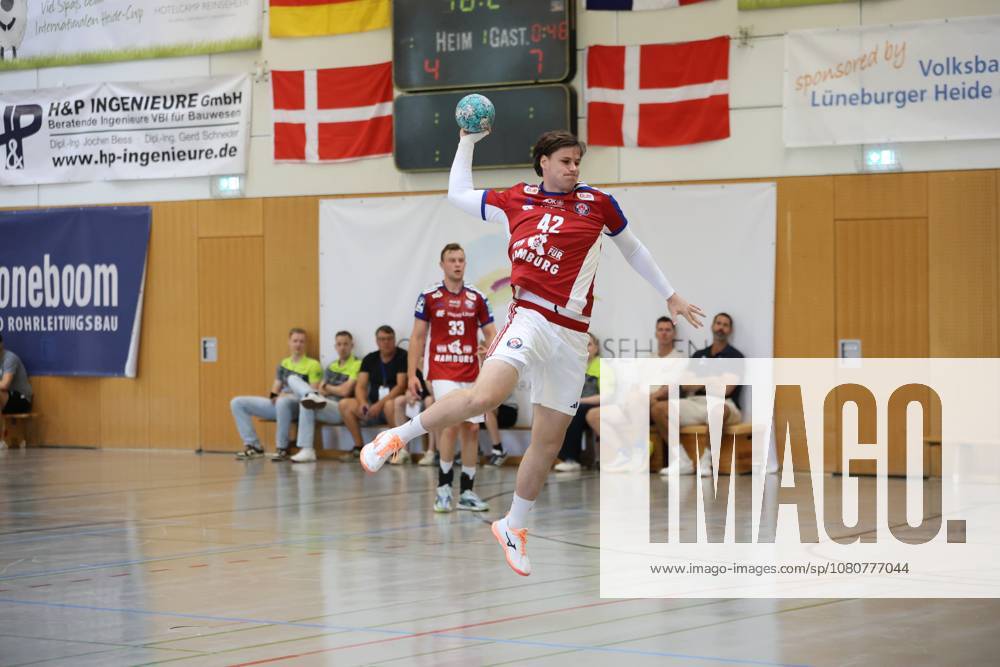 Jan Christian Schmidt, No. 42 of HSV Hamburg, jumps high and winds up for a shot; the ball is clearly visible in his raised hand. In the background, Moritz Sauter, No. 33 of HSV Hamburg, stands on the court, while other people sit on the bench. Jan Christian Schmidt, No. 42 of HSV Hamburg, takes a jump shot in Schneverdingen.