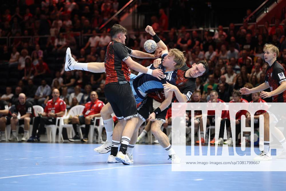 An HSV Hamburg player in a light blue and dark blue jersey drives past two MTV Braunschweig defenders with the ball in his hand. The matchup takes place in front of a packed crowd at the Volkswagen Halle in Braunschweig. The HSV Hamburg player fights his way through the MTV Braunschweig defense.