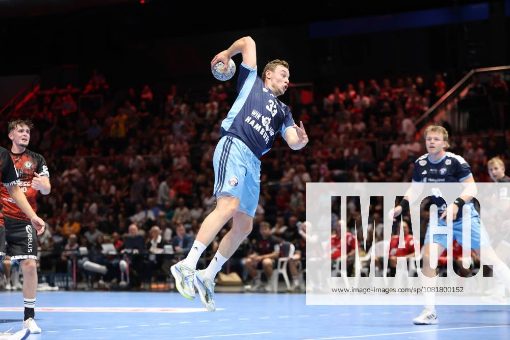 Moritz Sauter, No. 33 of HSV Hamburg, rises for a jump shot at the Volkswagen Halle in Braunschweig, holding the ball above his head, ready to shoot, while Martin Hovde, No. 81 of HSV Hamburg, watches the action in the background, while Tim Lietz of MTV Braunschweig watches from the left side of the frame. Moritz Sauter (33) of HSV Hamburg takes a jump shot in Braunschweig