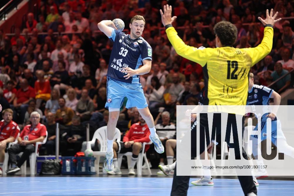 Moritz Sauter, No. 33 of HSV Hamburg, jumps and goes up for a shot, while goalkeeper Timon Serbest, No. 12 of MTV Braunschweig, spreads his arms. Another HSV player can be seen in the background. Moritz Sauter, No. 33 of HSV Hamburg, takes a jump shot against MTV Braunschweig.