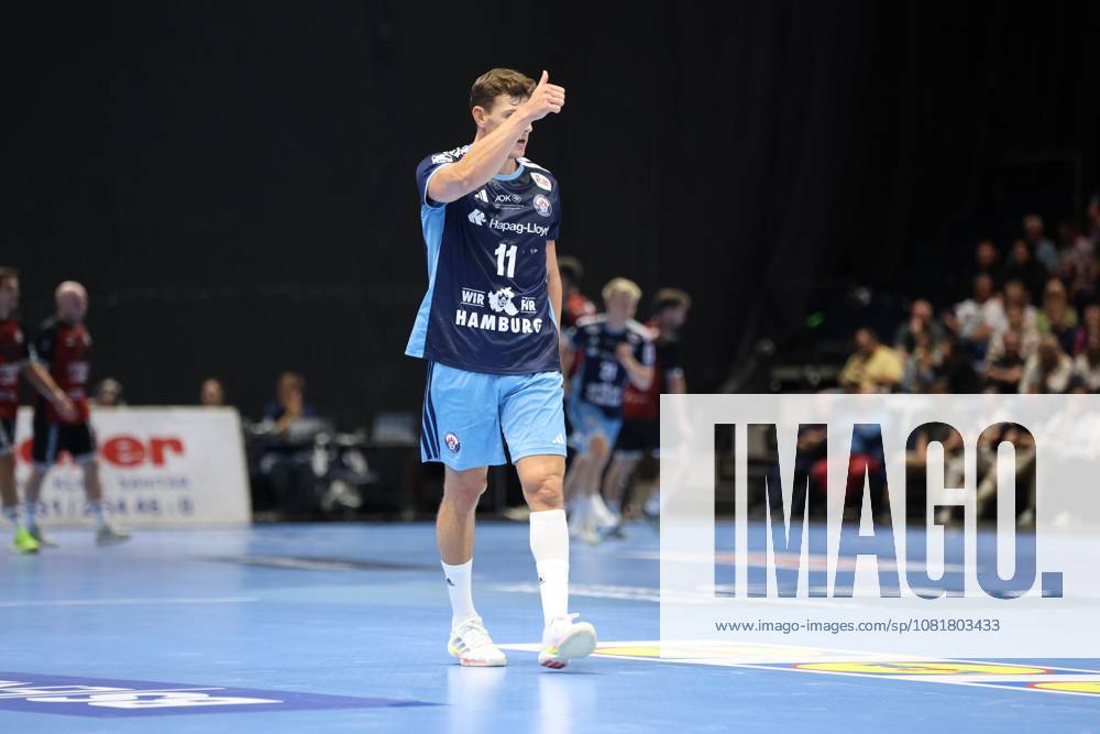 Nicolaj Jørgensen (No. 11, HSV Hamburg) stands on the court at the Volkswagen Halle in Braunschweig and gives a thumbs-up, raising his hand to his face. In the background, other players from both teams and spectators in the stands can be seen. Nicolaj Jørgensen (No. 11, HSV Hamburg) gives a thumbs-up.