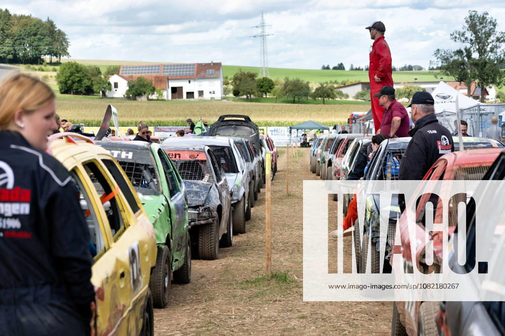 Numerous stock cars are lined up in two rows, ready for a race at the FUN CROSS Stock Car Race near Dietenwengen. Drivers and crew members wait among the heavily marked vehicles for the race to continue. FUN CROSS Stock Car Race in Eberhardzell Eberhardzell Dietenwengen