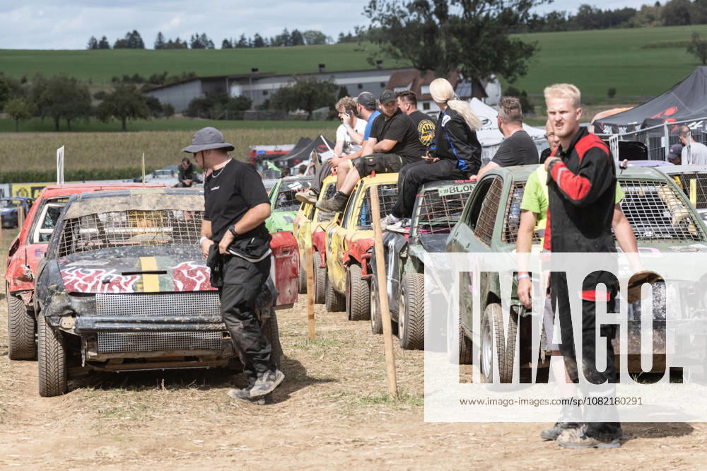 Drivers and crew members wait among numerous heavily battered stock cars in the paddock at the FUN CROSS stock car race near Dietenwengen as they await the next phase of the race. Several participants are sitting on the cars, watching the action. FUN CROSS stock car race in Eberhardzell. Eberhardzell, Dietenwengen
