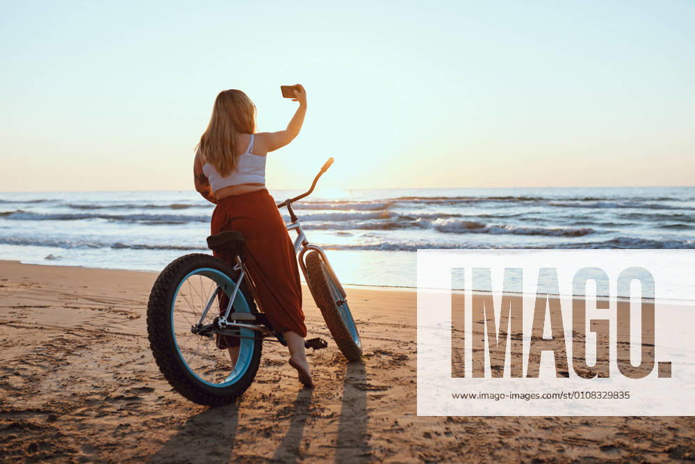 Cheerful chubby woman taking selfie on smartphone while resting with bicycle at sunny shore, Model released, Property released