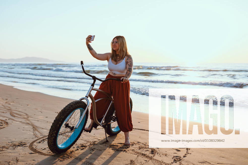 Cheerful chubby woman taking selfie on smartphone while resting with bicycle at sunny shore, Model released, Property released