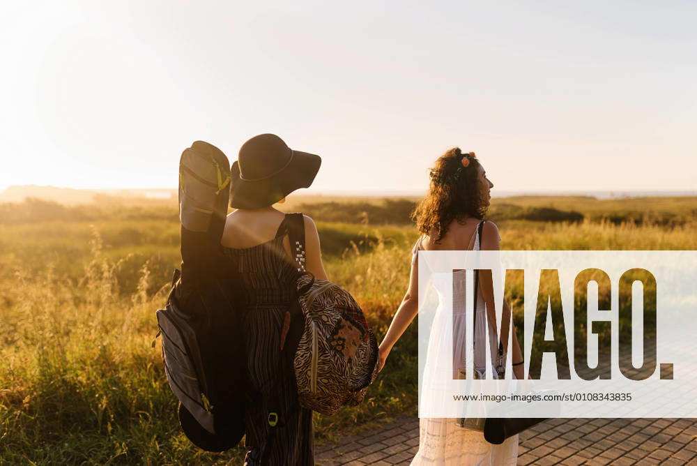 IMAGO Creative: Sommer, Abendspaziergang Back view of couple of girlfriends walking with musical instruments and holding hands while enjoying sunset in summer, Model released
