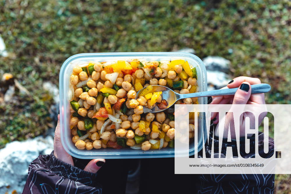 From above crop woman in warm jacket holding plastic container of chickpea with veggies having lunch on meadow while hiking in Spain, Model released