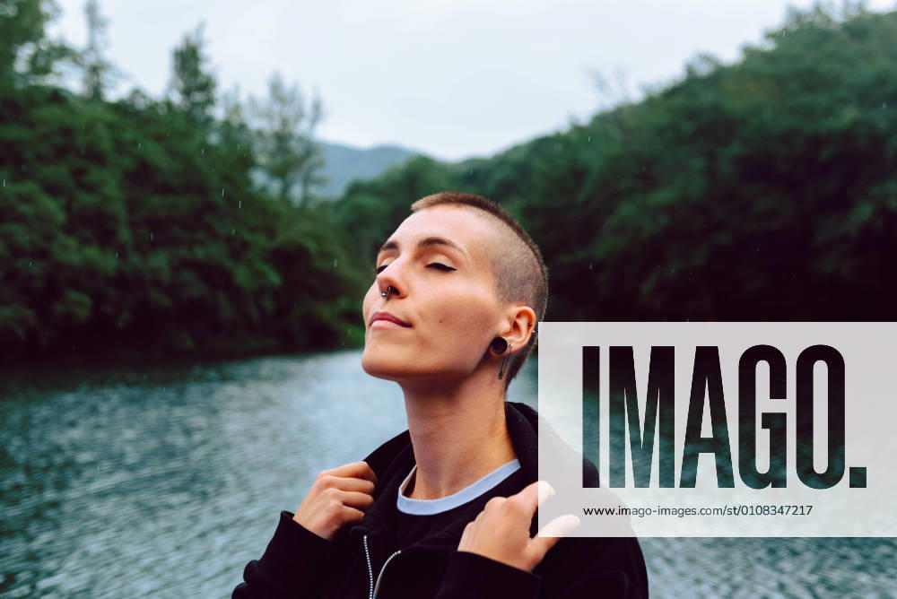 IMAGO Creative: Ende des Sommers Young extraordinary woman with short hairstyle and piercing in casual clothing facing head up with closed eyes to sky with pond among green plants on blurred background, Model released