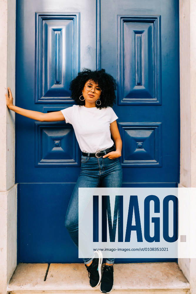 Pretty ethnic woman in white t-shirt and jeans leaning on blue door and looking at camera, Model released