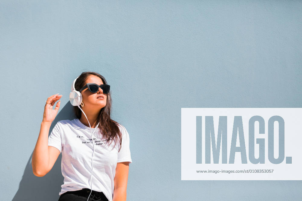 Carefree beautiful woman in casual outfit and headphones on neck standing with hands up beside blue wall of building on city street, Model released