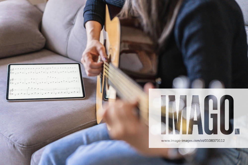 IMAGO Creative: Bildung, Musikerziehung Woman playing guitar sitting on her couch at home and learning with online lessons with a digital tablet with a digital tablet with a blank screen from above, Model released, Property released