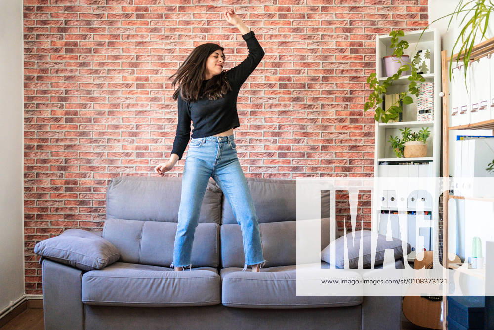 Young woman dancing on a couch in her house. Behind it is a brick wall, Model released, Property released