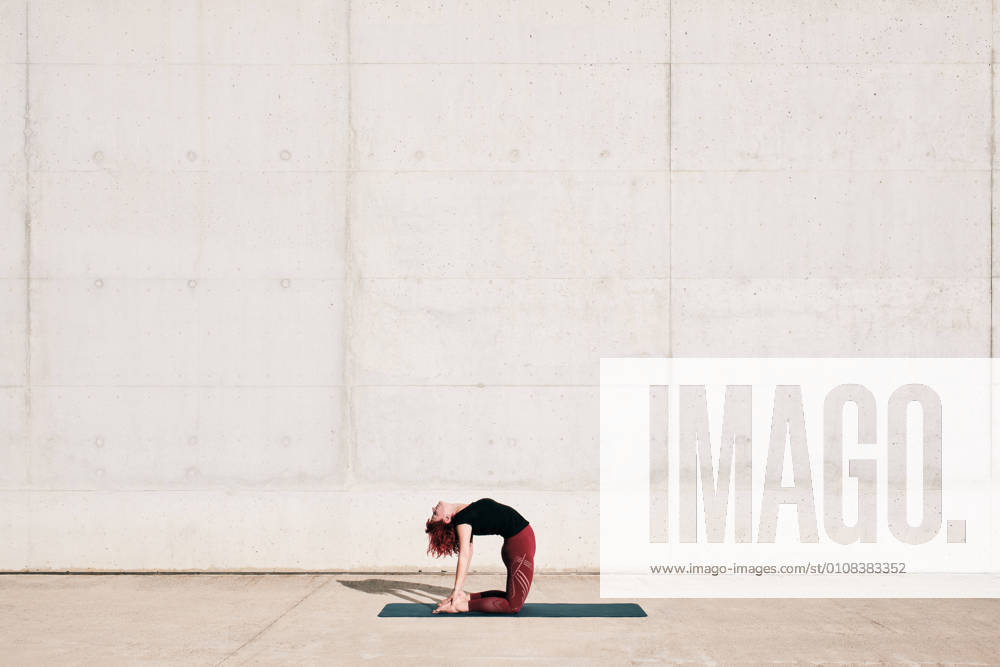 IMAGO Creative, Urban Lifestyle Trendy fit woman in sportswear doing yoga in camel pose on sports mat alone on street against concrete wall in sunny day