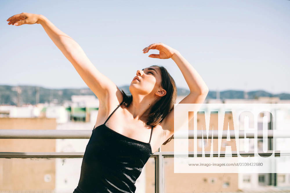Graceful female dancer in moment of performing element with outstretches arms looking up on summer terrace on background of palm trees