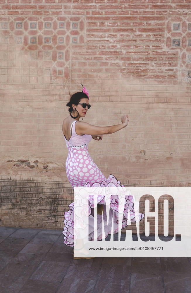 Side view of eccentric cheerful woman in colorful pink costume smiling and dancing by brick wall on sunny day