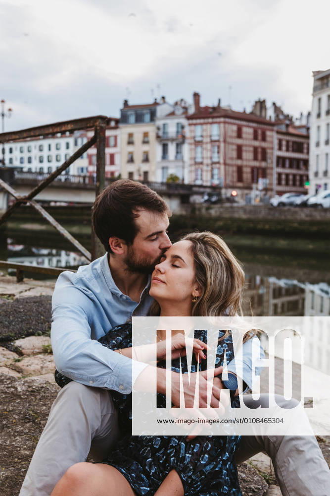 Positive young couple in casual clothes enjoying romantic date while sitting together on stone border in city with old buildings in background