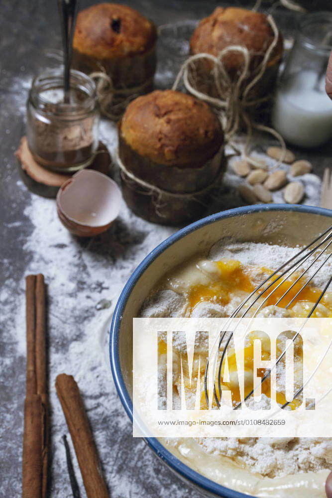 Housewife with whisk mixing products in ceramic bowl while preparing dough for typical Christmas panettone cake on table with baked panettone and ingredients