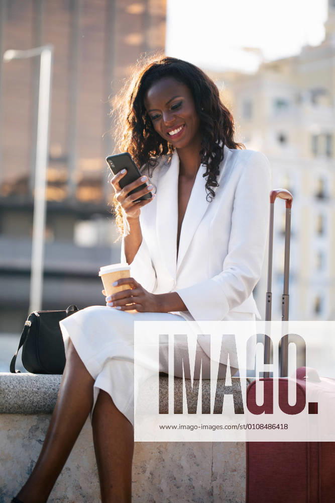 Positive African American businesswoman in elegant white clothes browsing cellphone and having coffee while sitting on street with suitcase, Model released, Property released