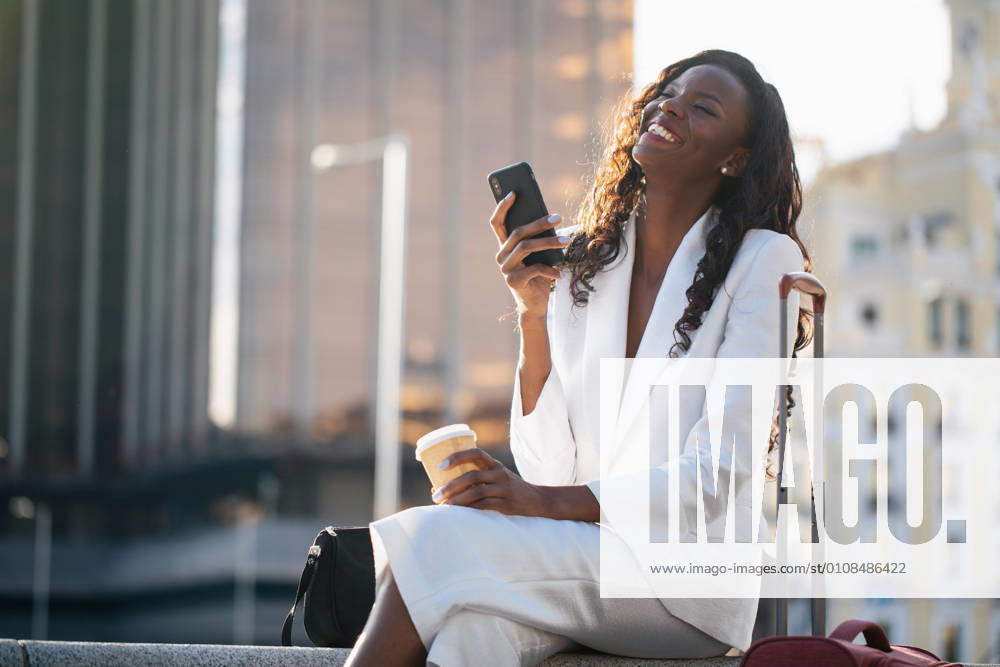 Positive African American businesswoman in elegant white clothes browsing cellphone and having coffee while sitting on street with suitcase, Model released, Property released