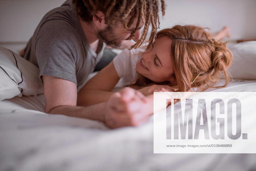 Tender man with dreadlocks hugging and kissing woman with red hair while lying on stomach on bed together and relaxing on weekend