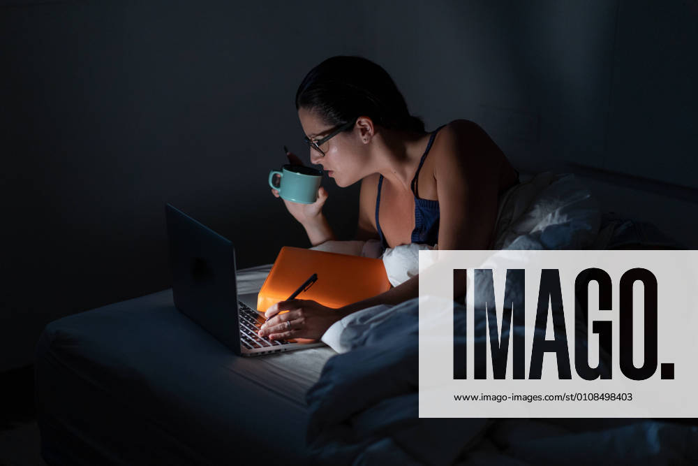 Focused female entrepreneur lying on bed and browsing laptop while drinking coffee and working remotely at night