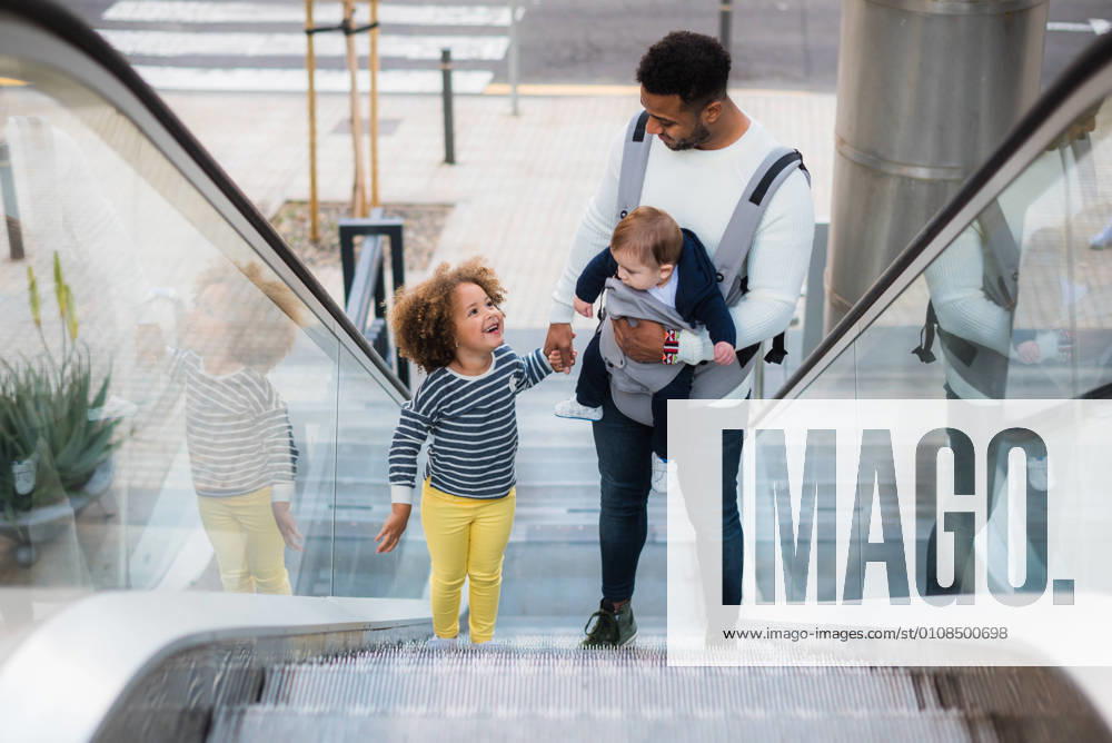 From above of positive young ethnic man carrying infant son and holding hand of cheerful little daughter while standing on escalator in city, Model released